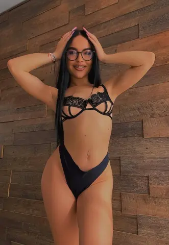 Sasha_Lorrenz poses with her hands in her long black hair, wearing black lace lingerie and glasses against a wood-paneled wall.