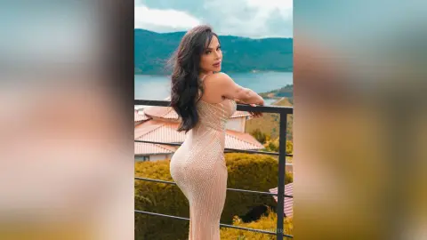RoxannaRivera poses overlooking a lake in a tight, sparkling gold dress that hugs her curves, showing her back and long dark hair.