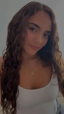 Young woman with long curly brown hair wearing a white tank top and gold oval pendant necklace.