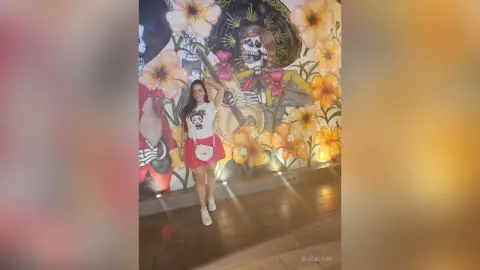 Smiling woman in a white t-shirt and red skirt posing in front of a colorful Dia de los Muertos mural.