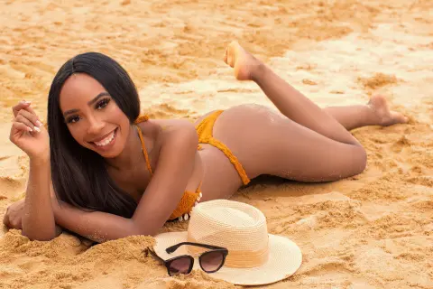 Smiling Black woman in a yellow bikini relaxing on a sunny sandy beach with a straw hat and sunglasses.