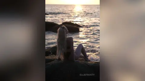 EllieFrosty sits naked on a dark rock overlooking the ocean during a sunset.