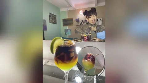 Young woman with tattoos in a blue top enjoying a colorful tropical cocktail in a bright, modern room.