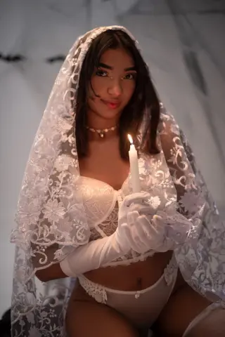 Ethereal woman in white lace lingerie and veil holding a candle in a moody, romantic setting.