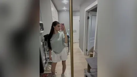 Young woman in a sage green t-shirt and white shorts taking a mirror selfie in a bright modern hallway.