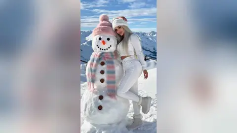 Blonde woman in white winter outfit posing with a large snowman in a sunny snowy mountain landscape.