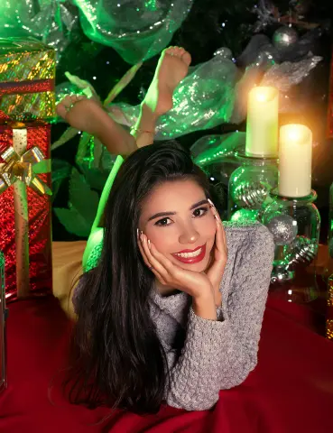 Smiling woman in a grey sweater relaxing near a glowing Christmas tree and festive holiday decorations.