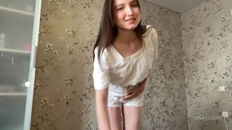 Young woman in a white blouse and denim shorts smiling in a room with floral wallpaper.