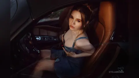StelaBrown sits in a brown leather car seat wearing a blue denim dress that exposes her cleavage.