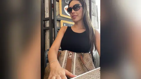 Stylish woman in a black tank top and sunglasses sitting at an outdoor cafe with a designer Guess handbag.