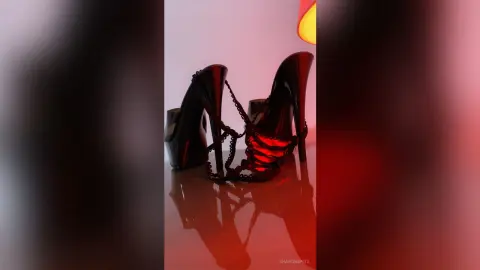 Elegant black patent leather high heel platform sandals with ankle straps on a reflective surface under red lighting.