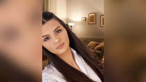 Young woman with long dark hair wearing a white shirt in a cozy, warm-lit living room.