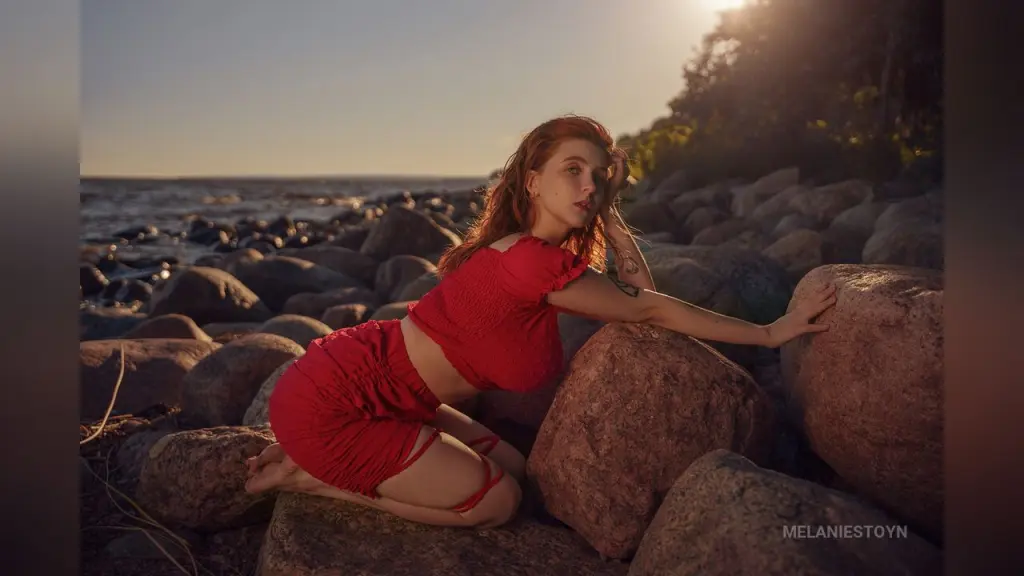 MelanieStoyn โ Redheaded woman in a red summer outfit sitting on coastal rocks by the ocean at sunset.