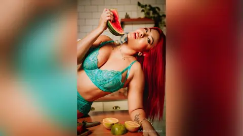 IsabellaEstrada poses in a teal lace bra and panties, holding a slice of watermelon near her face with her long red hair down.
