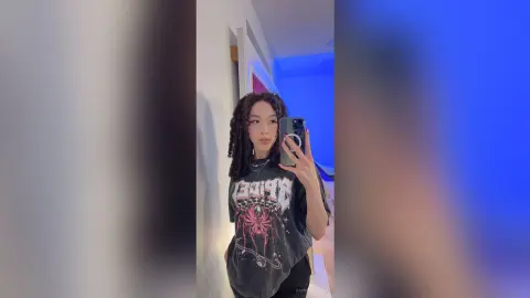Young woman with dreadlocks in a graphic black t-shirt taking a moody mirror selfie in a blue lit room.