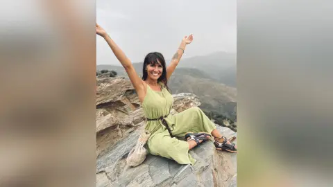 Smiling woman in a sage green linen jumpsuit sitting on mountain rocks under a cloudy sky.