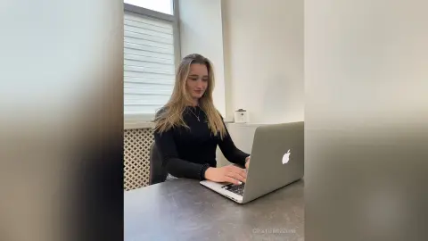 Young woman in a black long-sleeve shirt working on a laptop at a desk in a bright, modern office setting.