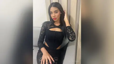 Glamorous woman in a black lace long-sleeve top sitting on a grey sofa with a confident expression.