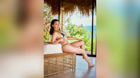 IsaReed sits on a white furry bench in a blue bikini, posing outdoors with a tropical view behind her.