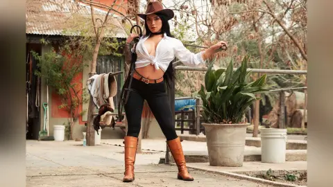 FlorenciaCopper stands outdoors in a white button-down shirt and black leggings, wearing a brown cowboy hat and boots while holding a riding crop.