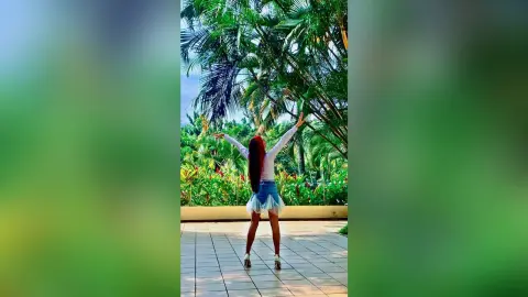 Cheerful woman in a white long-sleeve top and denim skirt raising arms in a lush tropical garden setting.