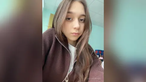 Young woman with long brown hair wearing a brown zip-up sweater in a bright indoor setting with a calm mood.