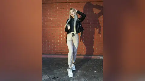 Stylish blonde woman in a black leather jacket and beige joggers posing against a red brick wall at night.