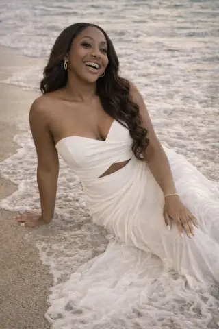 Smiling woman in a white strapless gown sitting on a sandy beach by the ocean waves.