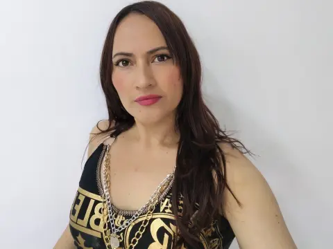 Woman with long brown hair wearing a black graphic tank top and layered silver necklaces against a white wall.
