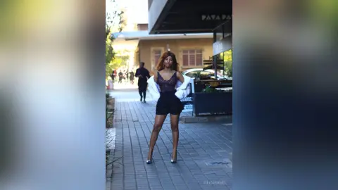 Stylish woman in a black lace top and shorts walking on a city sidewalk during a sunny day.