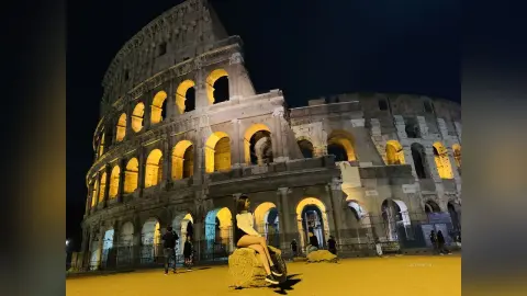 LucyMartin sits on a stone ledge in front of the illuminated Colosseum at night wearing a white top and light-colored skirt.