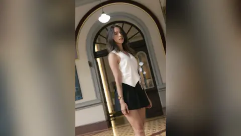 ValentinaGarcez poses in a white sleeveless top and black skirt, standing in a room with a large arched doorway and a hanging light bulb.
