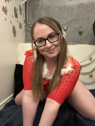Smiling woman in a red festive outfit with white faux fur trim sitting on a bed in a cozy room.