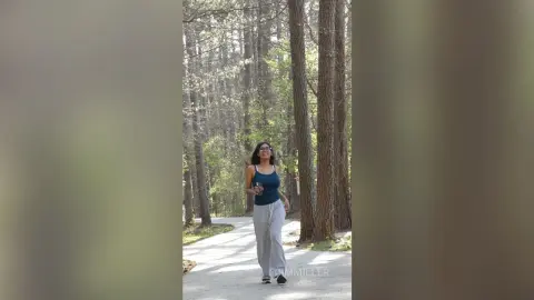 Smiling woman in a navy tank top and grey joggers walking on a sunny forest path through tall trees.