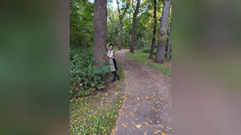 Young woman in a grey dress leaning against a tree on a peaceful autumn forest path covered in fallen leaves.