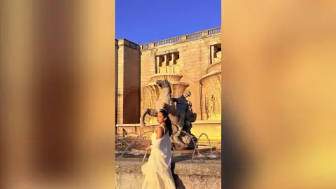 Woman in a white summer dress standing by a classical stone fountain at a sunlit historic palace garden.