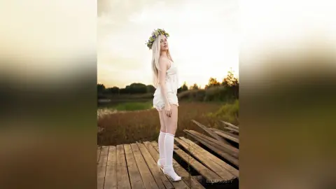 Blonde woman in a white lace outfit and flower crown standing on a wooden pier at sunset in a dreamy meadow.