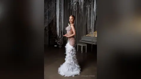StellaFletcher stands in a white feathered gown against a backdrop of hanging silver tinsel or fringe.