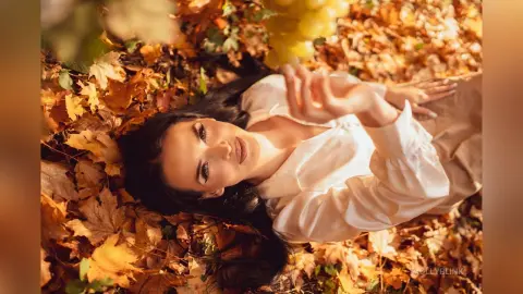 Elegant woman in a white blouse lying in golden autumn leaves for a dreamy fall fashion photoshoot.