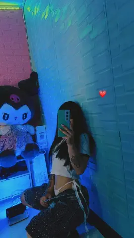 Young woman in a white crop top and patterned pants taking a mirror selfie in moody blue lighting.
