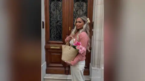 Blonde woman in a pink cardigan holding a straw bag and pink flowers in front of a vintage wooden door.