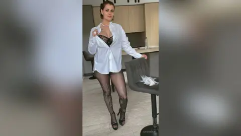 Elegant woman in a white button-down shirt and sheer black tights posing in a modern minimalist kitchen.