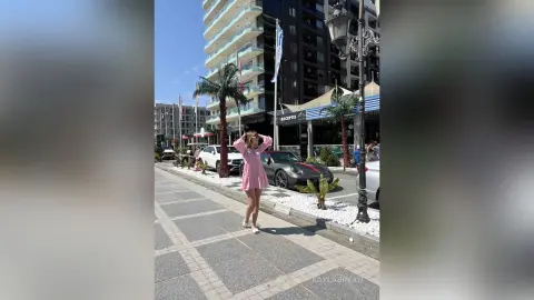 KaylaBryan poses in a short pink dress on a sunny city sidewalk next to a parked car.