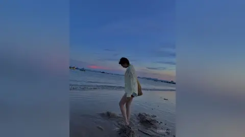 Young woman in a white linen shirt walking on a serene beach at sunset under a blue twilight sky.