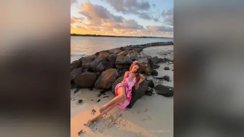 Young woman in a pink summer dress relaxing on a rocky beach at sunset under a cloudy sky.