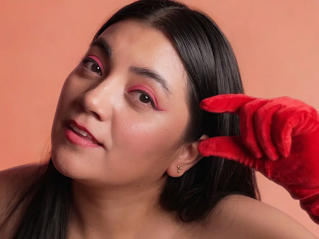 Woman with pink eye makeup and long dark hair posing against a peach background in a glamorous mood.