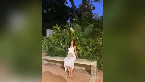 Woman in a white summer dress sitting on a stone bench in a lush tropical garden at twilight.