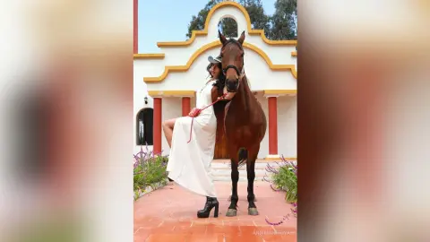 Elegant woman in a white maxi dress standing next to a brown horse in a sunny Mediterranean courtyard.