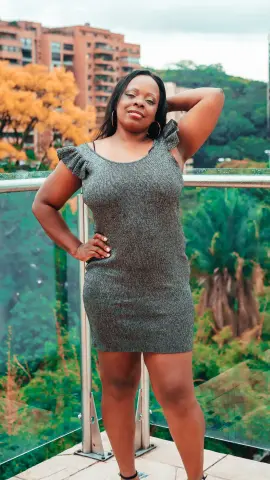 Smiling woman in a grey sleeveless mini dress posing on a balcony overlooking lush green trees and city buildings.