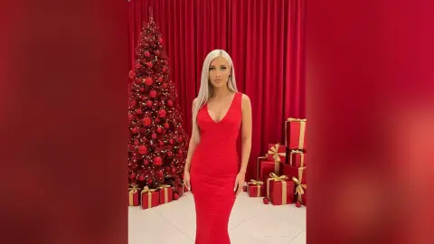 Elegant woman in a red evening gown posing by a decorated Christmas tree in a festive red room.